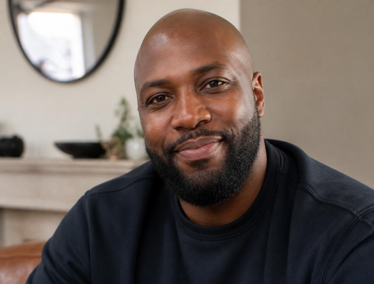 A bald Black man in his thirties with a full beard, smiling in a dark crewneck