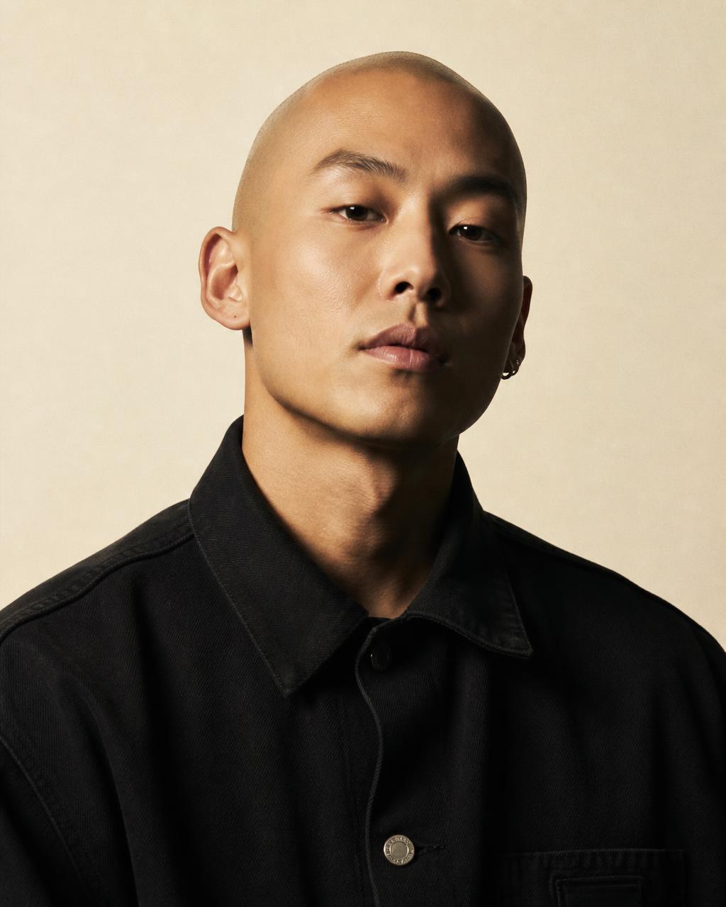 A young man with a freshly shaved head in a black workwear jacket, looking at the camera